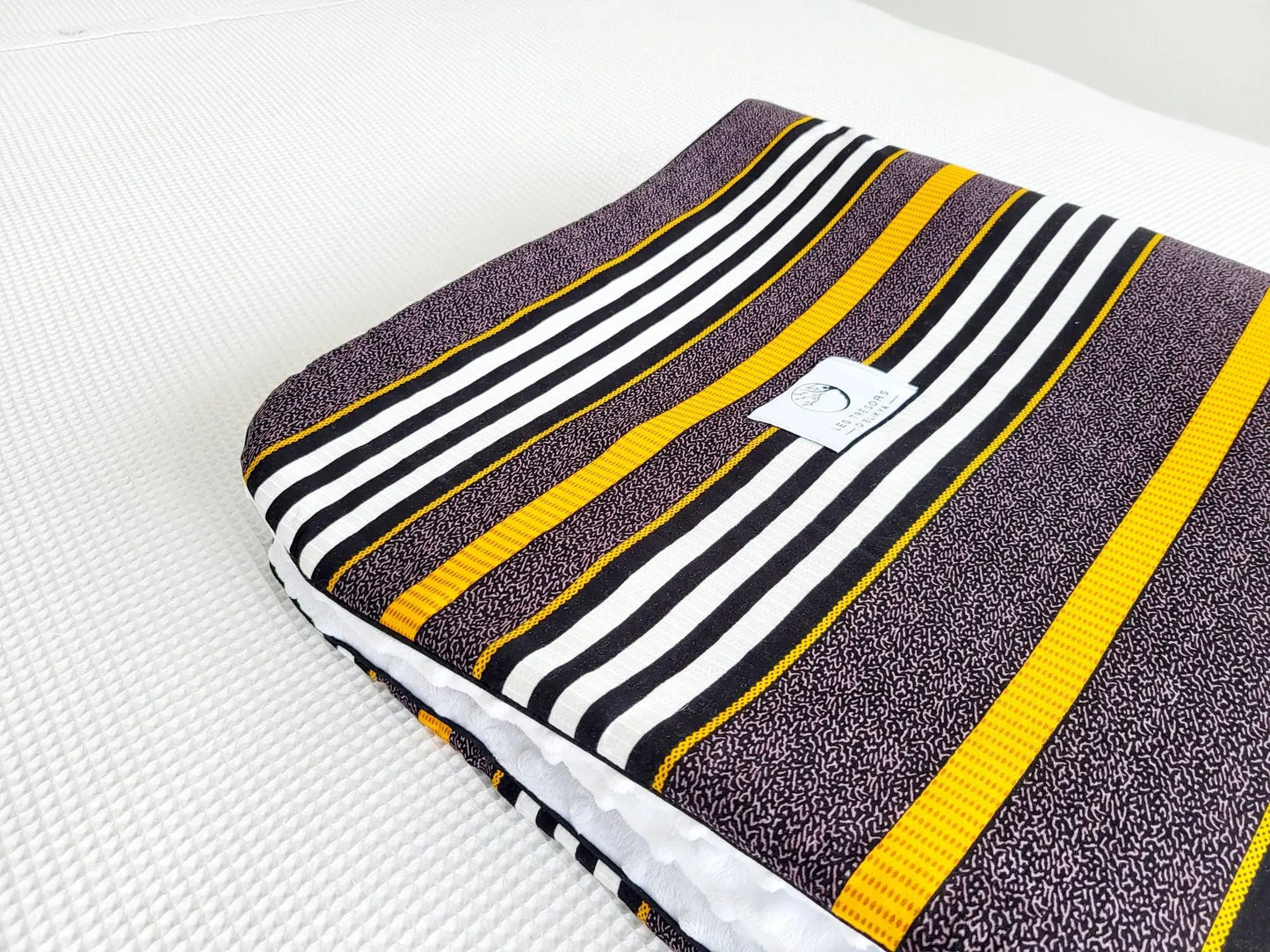 Zoom sur le carré de la couverture TIKA – tissu wax africain aux aux lignes raffinées mêlant des tons ocre, noir et blanc, dans une composition à la fois élégante et structurée, doublure minky ultra douce et finitions passepoils élégantes. Création artisanale Les Trésors d’Elikya pour envelopper bébé de douceur.