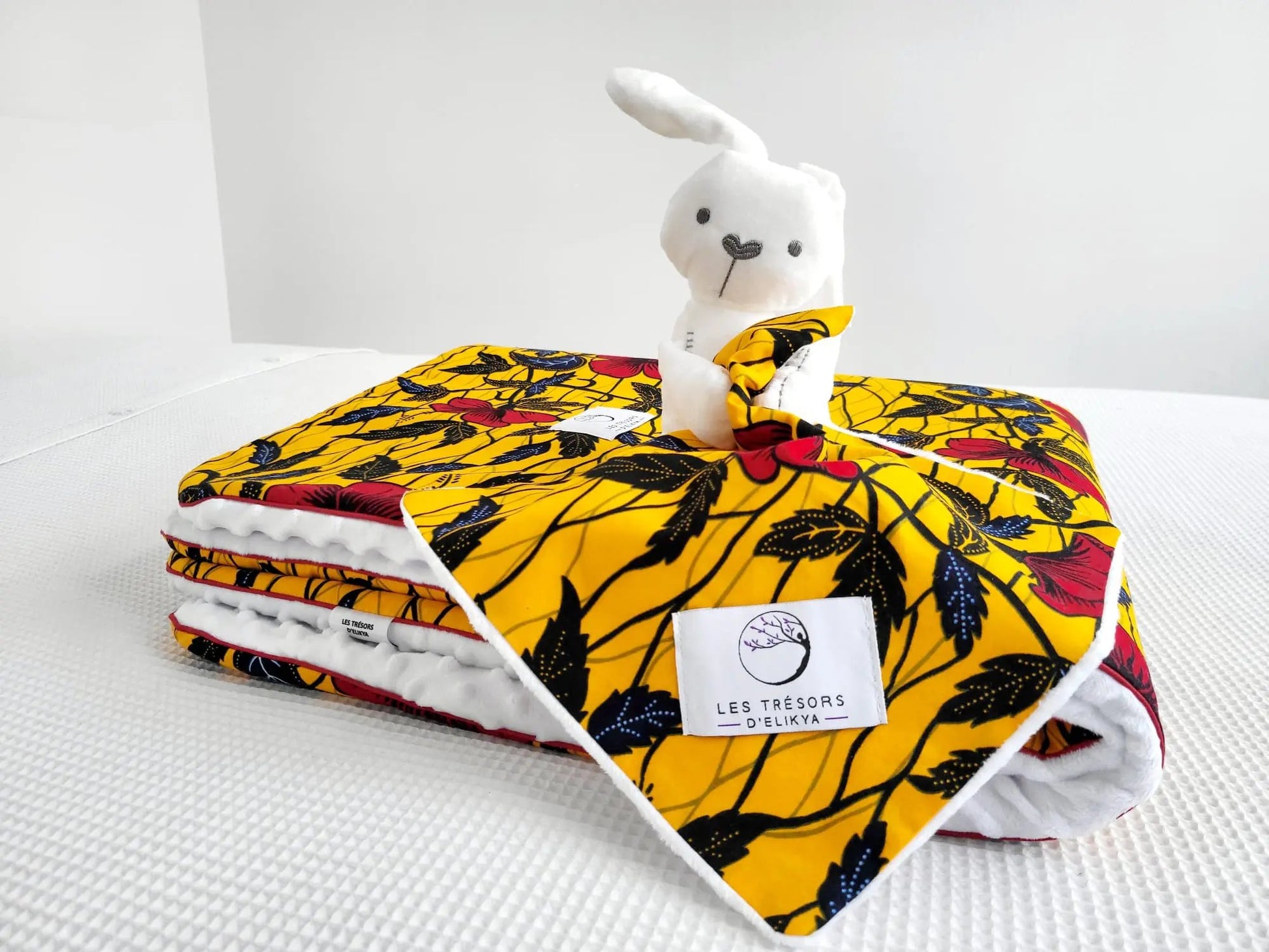 Zoom sur la mini couverture du doudou Bunny Wax AYANA et l’étiquette Les Trésors d’Elikya – peluche bébé avec sa mini couverture faite main en tissu wax africain aux motifs floraux. Détail de confection artisanale et idée de cadeau de naissance unique.