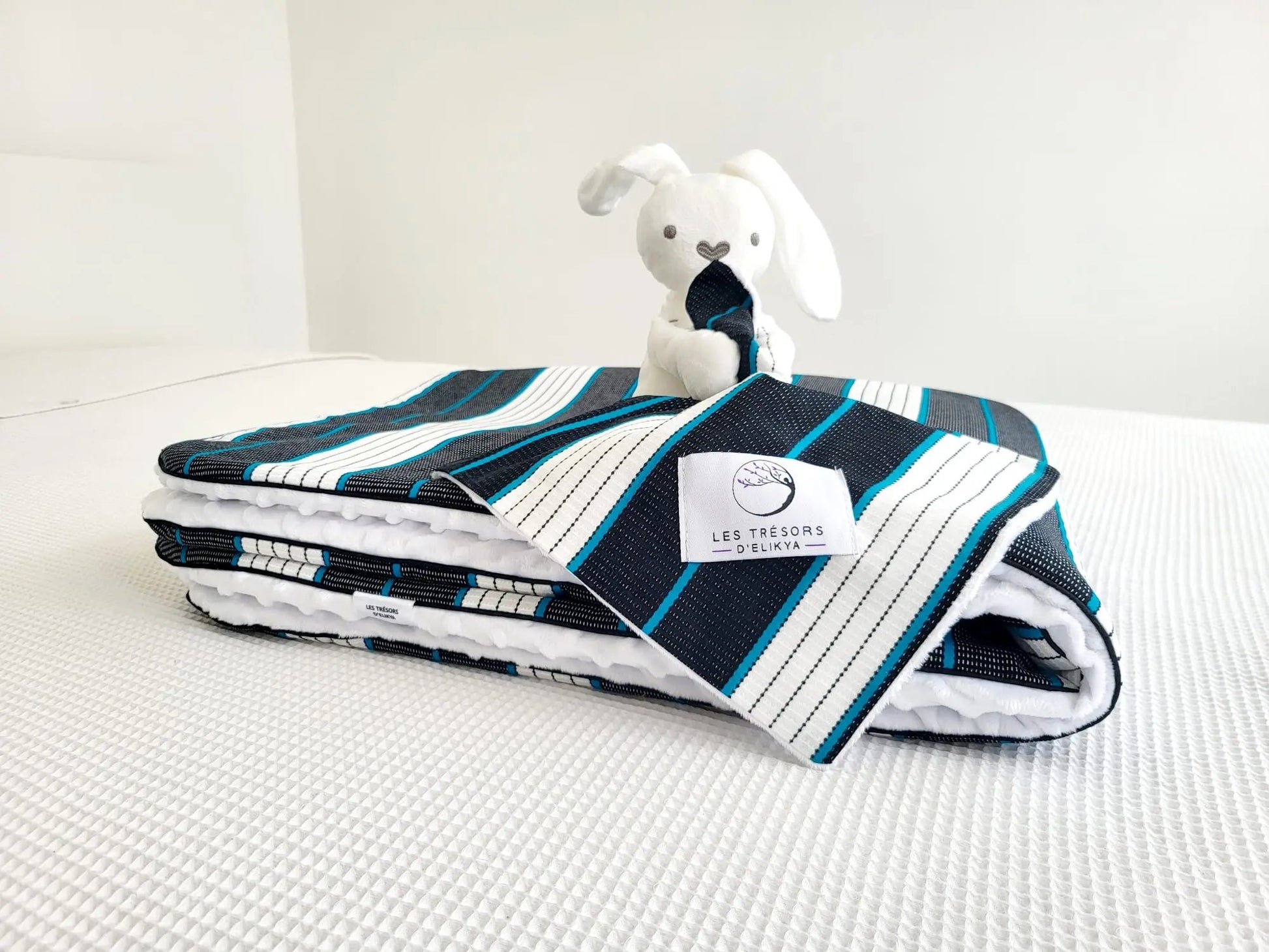 Zoom sur la mini couverture du doudou Bunny Wax KOBA et l’étiquette Les Trésors d’Elikya – peluche bébé avec sa mini couverture faite main en tissu wax aux lignes turquoise délicatement tracées, dans une danse équilibrée de noir profond et de blanc éclatant. Détail de confection artisanale et idée de cadeau de naissance unique.