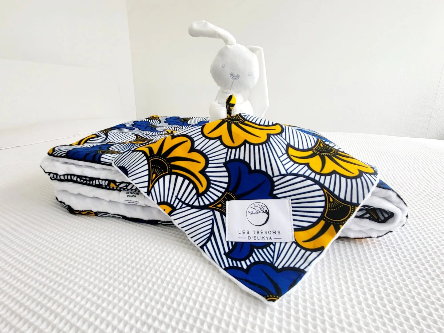 Zoom sur la mini couverture du doudou Bunny Wax MIRELYS et l’étiquette Les Trésors d’Elikya – peluche bébé avec sa mini couverture faite main en tissu wax africain fleurs de mariage jaunes et bleues sur fond blanc. Détail de confection artisanale et idée de cadeau de naissance unique.