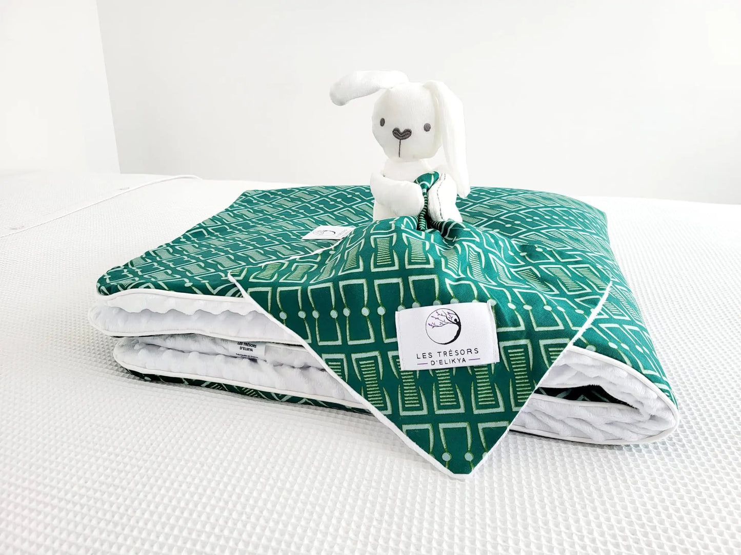 Zoom sur la mini couverture du doudou Bunny Wax NAIORIA et l’étiquette Les Trésors d’Elikya – peluche bébé avec sa mini couverture faite main en tissu wax africain avec un motif graphique vert profond orné de formes géométriques ivoire et de pois réguliers. Détail de confection artisanale et idée de cadeau de naissance unique.