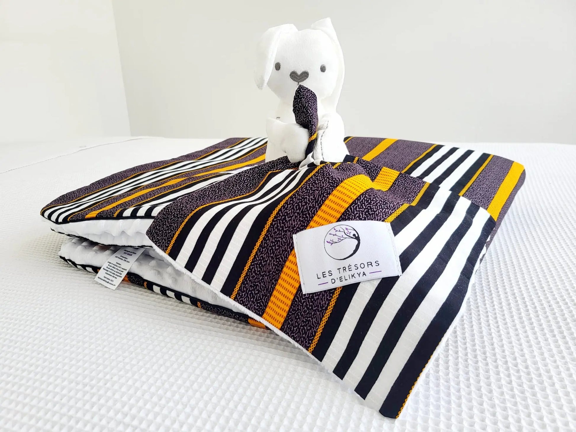 Zoom sur la mini couverture du doudou Bunny Wax TIKA et l’étiquette Les Trésors d’Elikya – peluche bébé avec sa mini couverture faite main en tissu wax africain aux lignes raffinées mêlant des tons ocre, noir et blanc, dans une composition à la fois élégante et structurée. Détail de confection artisanale et idée de cadeau de naissance unique.